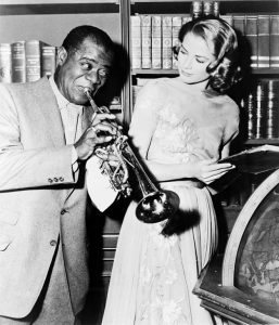 Louis "Satchmo" Armstrong blowing his trumpet while Grace Kelly looks on in a photograph from the set of the MGM motion picture "High Society." Louis Armstrong and Grace Kelly both featured along with Bing Crosby, Frank Sinatra and Celeste Holm.